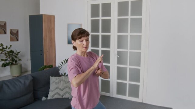 Medium shot of beautiful elderly Caucasian woman in pink t-shirt performing smooth, fluid arm movements while practicing tai chi in living room at home, maintaining flexibility in old age