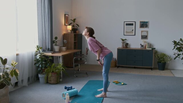 Wide shot of senior Caucasian woman in pink t-shirt, blue leggings doing forward body wave exercise while standing in living room at home, developing flexibility and strength in retirement