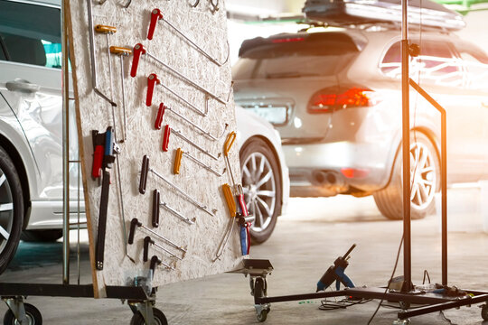 In a well-lit Garage, a rolling stand holds an assortment of automotive body repair tools. Two cars are parked in the background, one silver and one white, awaiting maintenance