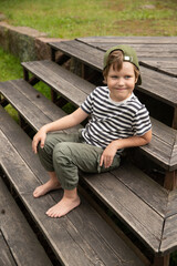 A boy sits on a wooden staircase outside in the summer. A boy laughs in a cap barefoot on a wooden platform near the lawn.