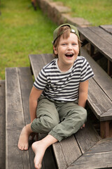 A boy sits on a wooden staircase outside in the summer. A boy laughs in a cap barefoot on a wooden platform near the lawn.