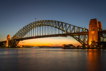 Fototapeta premium Iconic Sydney Harbour Bridge at twilight, beautifully lit with reflections on the water, under a golden sunset sky in Australia’s most famous harbor