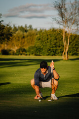 Obraz premium Smiling male golfer squats on the green to line up a putt, holding his club and preparing for a shot under golden evening sunlight on a scenic course.