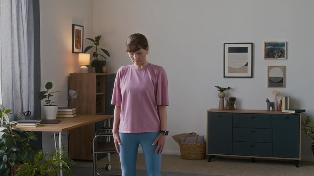 Medium shot of focused beautiful elderly Caucasian woman in pink t-shirt standing in living room at home, rotating head to side to warm up neck muscles and spine joints before beginning workout	