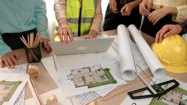 Architect and safety engineer discussion about building blueprint displaying on laptop on table with architectural document and wooden block scatter around at modern office. Closeup. Delineation.