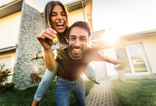 Happy young couple holding home keys after buying real estate - Husband and wife standing outside in front of their new house	 - Powered by Adobe