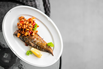 Grilled fish fillet with crispy skin topped with tomato vegetable salsa, garnished with parsley and lemon wedge, served on a white plate. Close-up of gourmet seafood dish