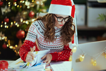 Christmas time. stylish 40 years old business woman in santa hat in eyeglasses in red Christmas sweater with documents and laptop working in modern green office with Christmas tree.
