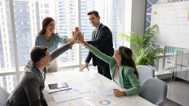 Diverse team of business professionals celebrate success with high-five gesture during corporate meeting in office, stacking their palms together as symbol of unity, express trust, finalize teamwork