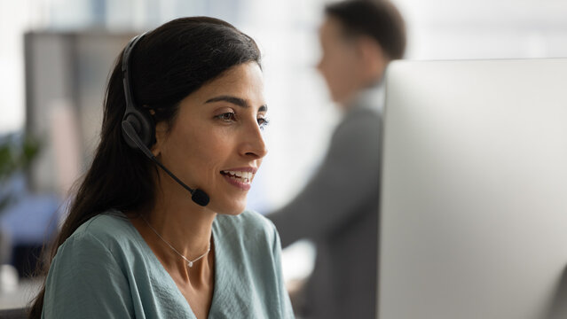 Close up of young Brazilian woman wearing headset attentively engaged in conversation with client, look at computer screen, working in professional customer service or technical support center office
