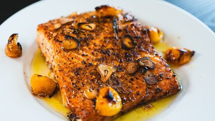 Succulent pan-seared salmon drizzled with olive oil, topped with crispy burnt garlic slices and roasted yellow tomatoes. Gourmet plating, highlighting textures and colors for food branding and culinar