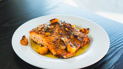 Succulent pan-seared salmon drizzled with olive oil, topped with crispy burnt garlic slices and roasted yellow tomatoes. Gourmet plating, highlighting textures and colors for food branding and culinar