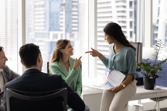 Group of happy colleagues gathered in modern high-rise office, focus on smiling woman holding business reports, points playfully at seated female colleague, enjoy friendly interaction during briefing