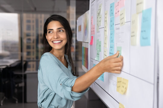 Smiling Latina professional woman placing sticky notes on large planning board, engaged in strategic planning, creative brainstorm session or workflow organization. Project management in modern office
