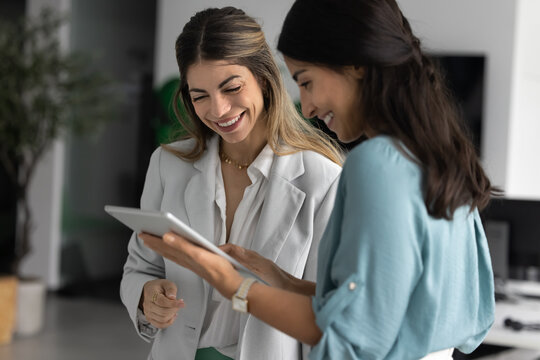 Two Latina colleagues reviewing project presentation on digital tablet in workplace, browsing through design mockups or marketing visuals, engaged in positive teamwork during workday using modern tech