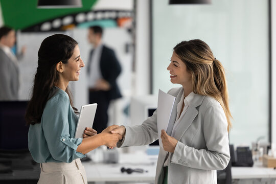 Two professional businesswomen shaking hands, greeting each other standing in modern office, making deal, concluding successful agreement or partnership, welcoming client, start business communication
