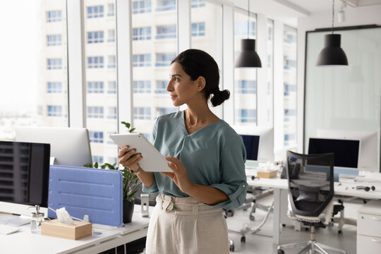 Pensive businesswoman working online using tablet standing in modern coworking space, looks thoughtfully away, reviewing data, reading report and considering strategic information displayed on device