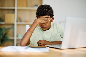 Portrait of teenage black boy using laptop computer and thinking and day dreaming looking through window at home. Teenage boy attending to online school class worried and tired with headache
