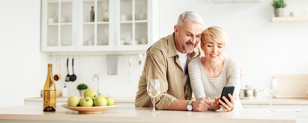 Online chat, call and communication with family remotely and celebrate at home during covid-19 quarantine. Smiling adult couple look at smartphone in kitchen interior with apples and glasses of wine
