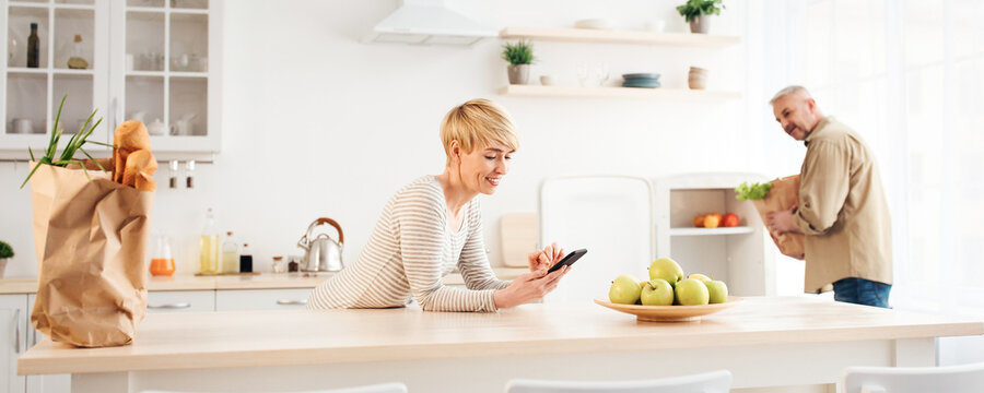 Service delivery of groceries during covid-19 lockdown, advice of modern food blogger. Cheerful senior man lays vegetables in refrigerator from paper bag, wife types in phone in kitchen interior