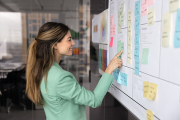 Hispanic woman participating in business planning session or project workflow review in modern workplace, points at section of board filled with sticky notes and structured documents. Task management