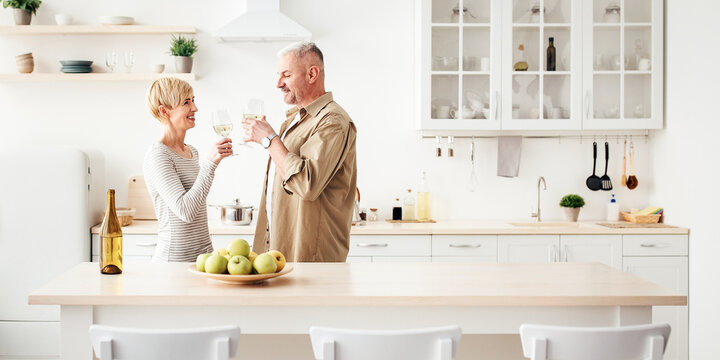 Romantic adult couple holding glasses, standing in modern cozy white kitchen interior. Cheerful married husband and wife drinking red wine, celebrate anniversary or holiday together at home, panorama - Powered by Adobe