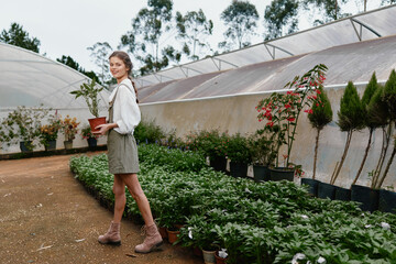 Young woman in a greenhouse holding a plant, portraying joy and connection with nature, dressed in a casual skirt and boots, amidst vibrant greenery
