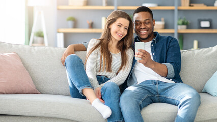 Weekend Pastime. Millennial Interracial Couple Relaxing With Smartphone At Home, Loving Black Man And White Woman Resting On Couch Together, Browsing Internet On Mobile Phone, Free Space