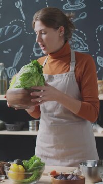 Vertical shot of female food blogger in apron list in ingredients and showing them to camera while filming cooking show at kitchen conter