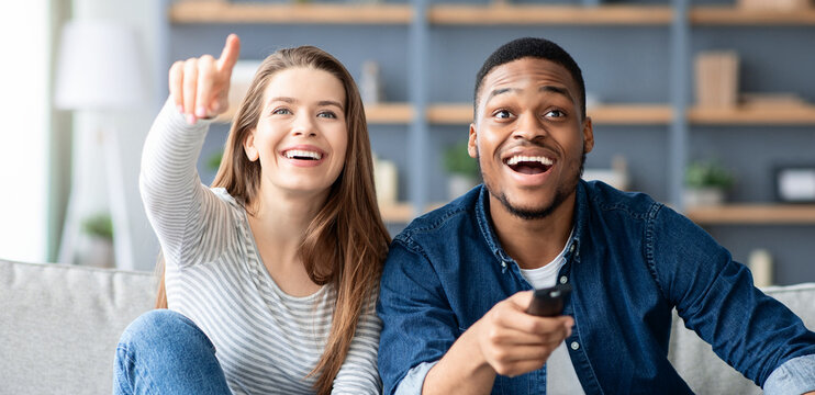 Cheerful multiracial couple having fun at home together, watching comedy movie on tv and laughing, excited interracial lovers sitting on couch in living room, woman pointing at television, closeup