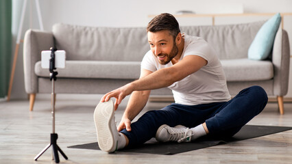 Cheerful sporty middle-aged man stretching on fitness mat, broadcasting with smartphone, having...