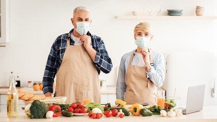 New normal, health care, be safety, modern food bloggers. Senior european man and wife in protective masks in modern kitchen interior with bright fresh vegetables on table and laptop, prepare meal