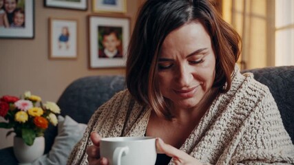 Cozy living room scene with a woman wrapped in a blanket, enjoying a hot drink. Warm lighting and framed photos in the background. Video shot at eye level.
