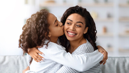 Loving african american school girl with curly hair kissing her beautiful mother, closeup portrait, copy space. Adorable black family young woman and her daughter bonding at home