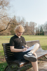A focused student works outdoors on a laptop and takes notes while sitting alone on a park bench on a sunny day