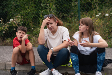 A mother is sitting with her two children outdoors; they appear frustrated, сhildren are arguing among themselves. Stressed mother sitting with challenging children outdoors in urban setting
