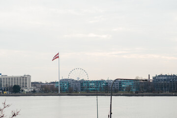 A calm riverside cityscape featuring a Latvian flag, a Ferris wheel, and modern buildings under a soft, overcast sky
