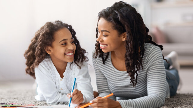 Joyful african american beautiful mother and daughter laying on floor and drawing together at home, using colorful pencils and coloring book, closeup. Art and craft, leisure activity concept