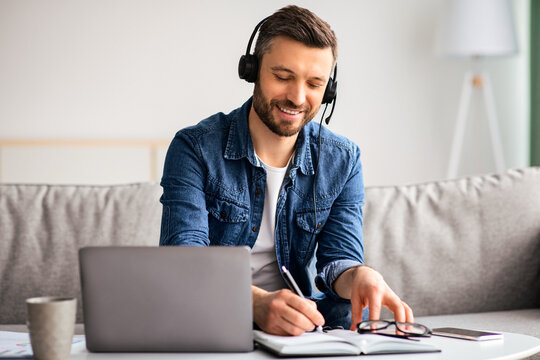Positive middle-aged man attending online training from home, sitting on couch in front of coffee table with laptop, using headset, taking notes, copy space. E-education, online class, training - Powered by Adobe