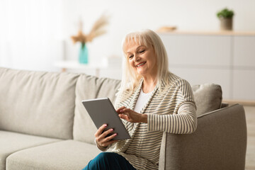Free Time Concept. Portrait of smiling senior woman holding digital tablet, reading news or e-book, sitting on comfy sofa. Happy mature lady browsing internet, watching video online, touching screen