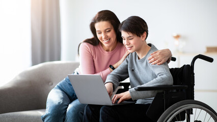 Distance education concept. Joyful disabled teenager in wheelchair using laptop, making homework...