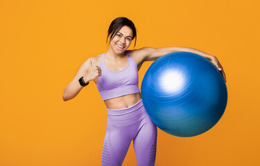 Fototapeta premium Young sporty black woman holding fitness ball and showing thumb up, standing on yellow background, studio portrait. Workout, sport, motivation concept