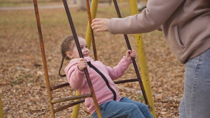 Obraz premium little kid swinging on a swing and laughing while flying up, an autumn playground, a happy family, a mother rolls child in city park, childhood dream to fly, smile at mom on a walk, play with a baby.