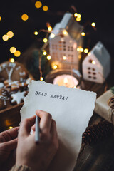 Christmas letter on craft paper to Santa Claus with text: Dear Santa. Cozy home interior with candles, lights, handmade toys, gingerbread cookies. Wishlist for presents from a child or adult