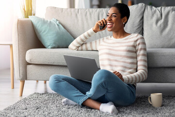 Fototapeta premium Distance Job Advantages. Portrait Of Cheerful Black Freelancer Lady Using Laptop Computer And Talking On Cellphone At Home, Happy African American Lady Enjoying Working Remotely, Free Space