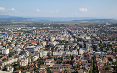 Fototapeta premium An elevated, panoramic shot of a city center with dense residential and commercial buildings under a clear sky. In the background, rolling hills and a mountain range can be seen