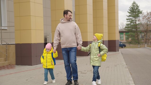school classes, father leads little children with backpacks holding hands, happy family, dad walks with kid around school yard, accompany lessons study, preschool education baby, school bag shoulders.