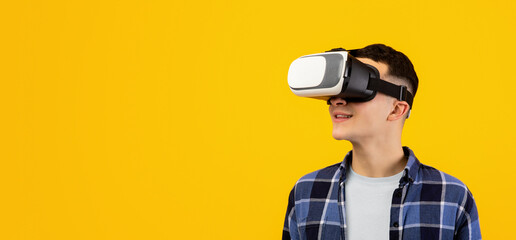 Cool young guy wearing VR headset, exploring cyberspace, using virtual reality for entertainment or studies on orange studio background. Funky millennial man experiencing futuristic technologies