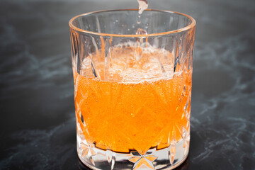 A crystal glass overflows with a refreshing orange beverage, featuring bubbles and ice, set against...