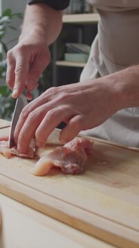 Vertical tilt-down shot of male chef expertly preparing chicken breasts, slicing them with knife in his kichen